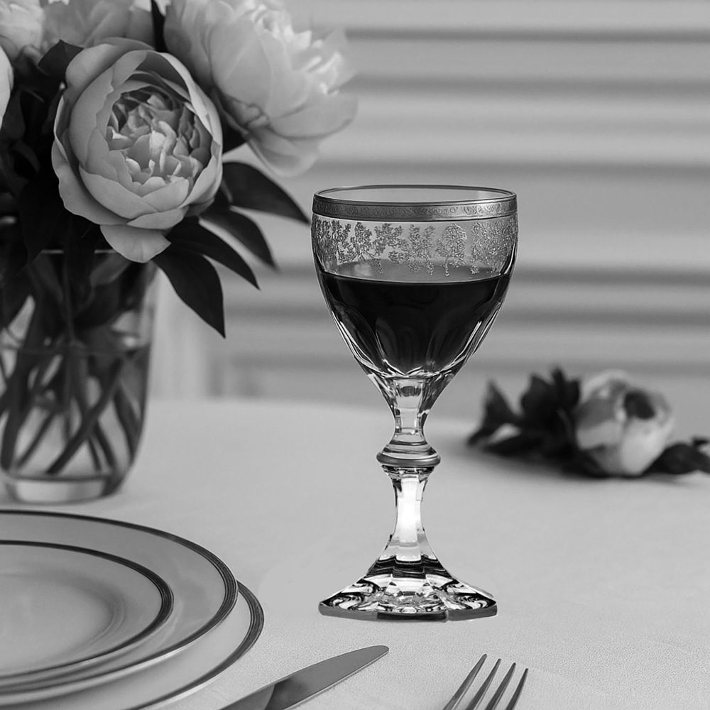 Elegantes Kristall-Rotweinglas mit dunklem Wein auf hellem Tisch. Rand und Schale mit floralem Muster verziert, schlanker Stiel und facettierter Fuß. Links im Hintergrund Blumenstrauß in Vase; vorn Teller und Besteck.
