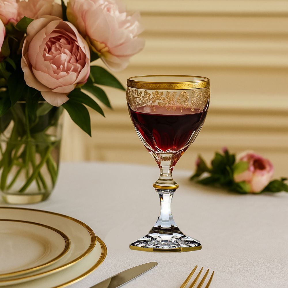 Elegantes Kristall-Rotweinglas mit rotem Wein auf weißem Tisch. Goldene, filigrane Ornamente am Rand und Fuß. Im Hintergrund rosa Pfingstrosen in einer Vase sowie eine festlich gedeckte Tafel mit Tellern und Besteck.